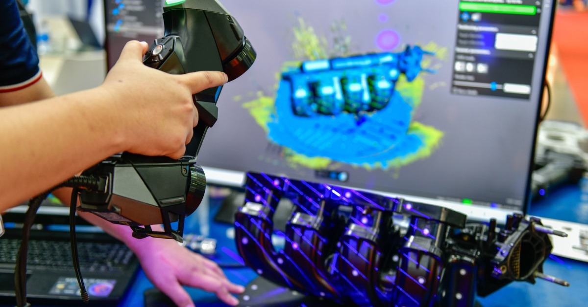 Someone using a 3D scanner to scan an engine motor into a computer. The 3D rendering is on the screen behind it.