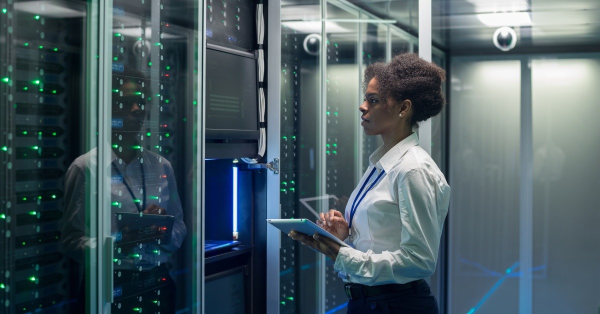A woman with a tablet looking at a large group of data servers. She has one of the glass doors of the wall open.