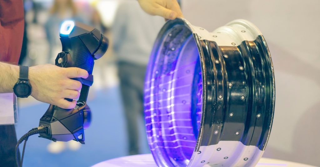 Someone using a 3D scanner on a metal car rim. There are purple lines coming from the scanner on the front of the wheel.