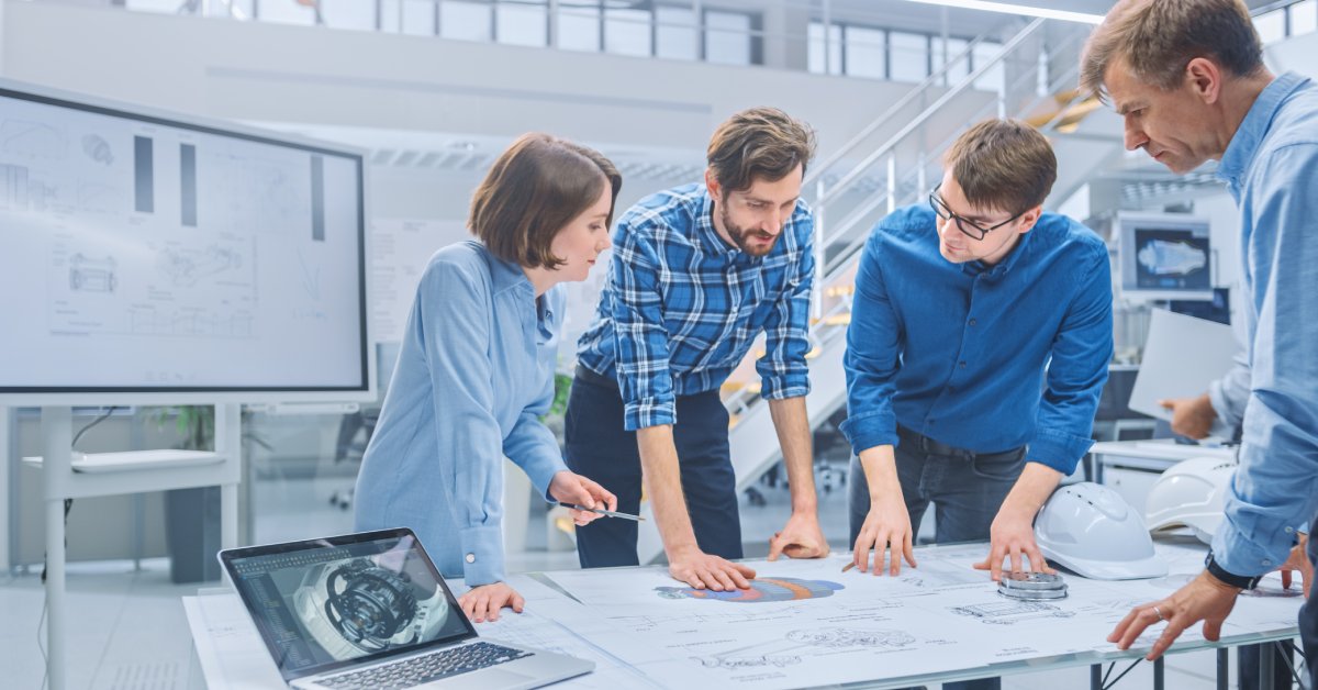 A group of people standing around a table as they look at product designs laid out on the table. They're in a modern, well-lit office with white walls.