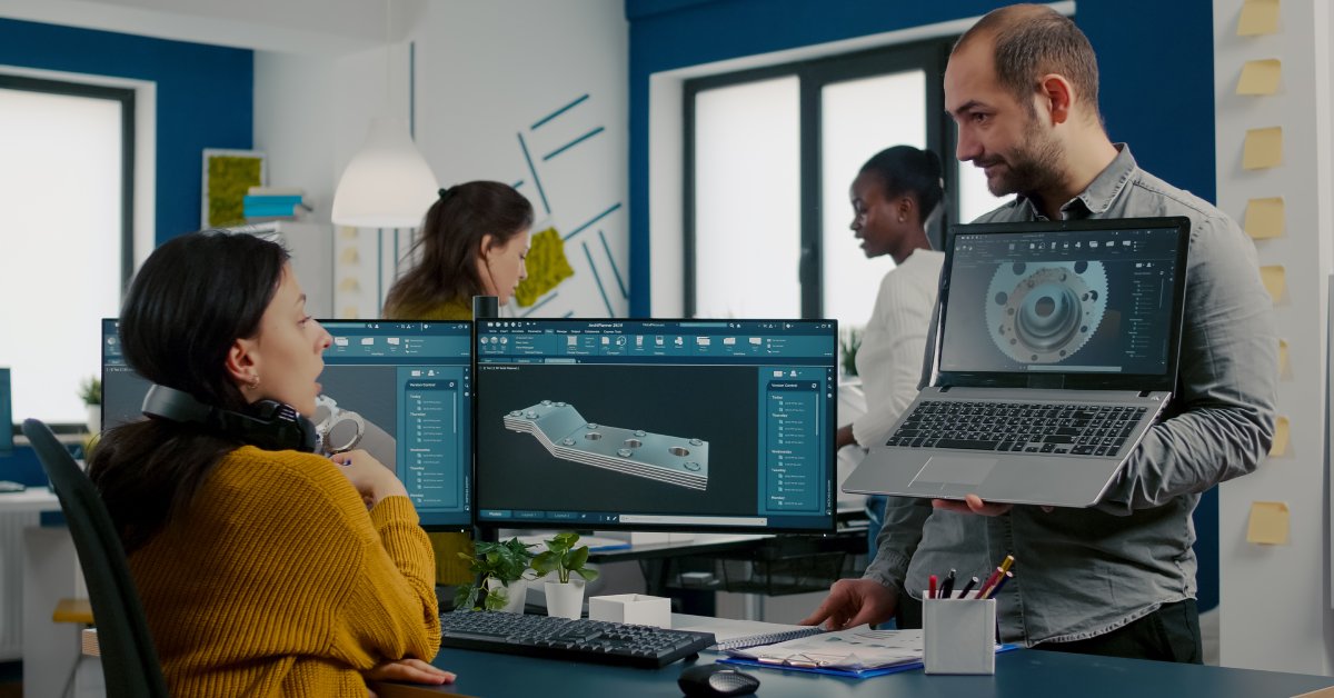 A woman sitting at a desk with two screens displaying product parts. She's looking over at another man's laptop that has a 3rd product part.