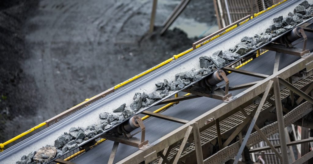 A long line of black rocks moving down a conveyor belt. The belt is high up in the air with a road passing underneath it.