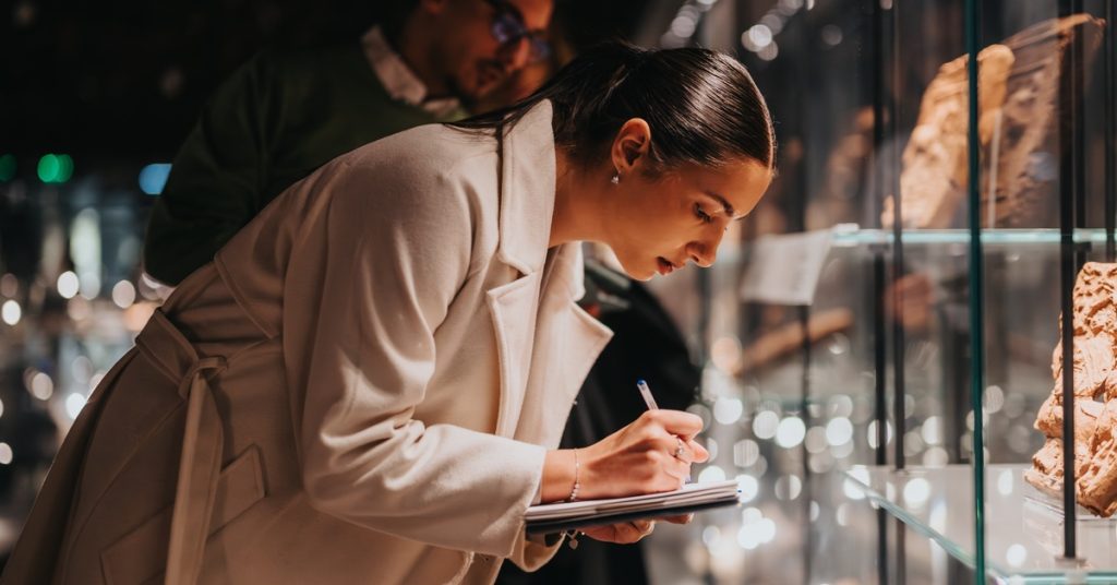 A woman bending over to look at an ancient artifact. She is writing some notes down in her journal.