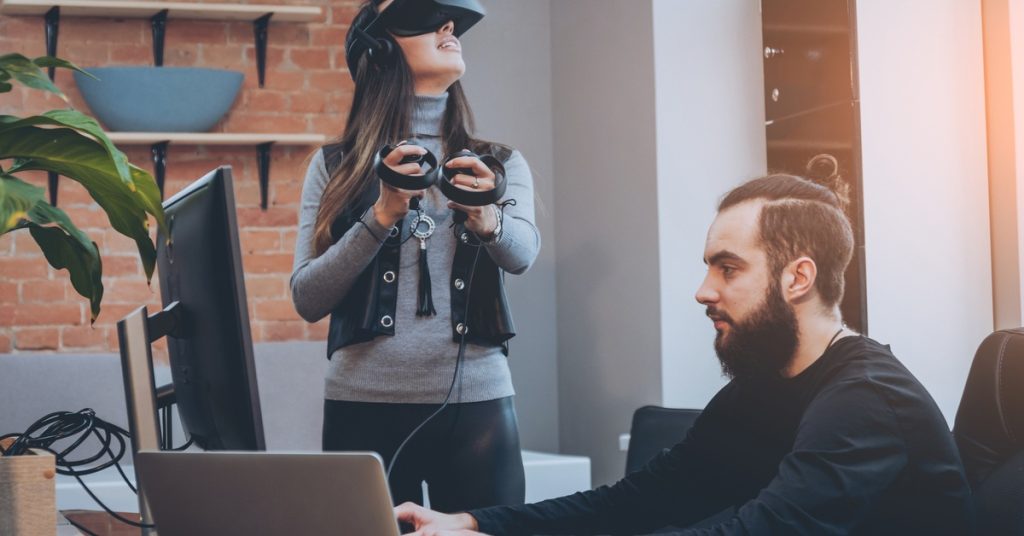 A woman using a VR headset while a guy sits at a desk, working on a computer. She has VR remotes in both hands.