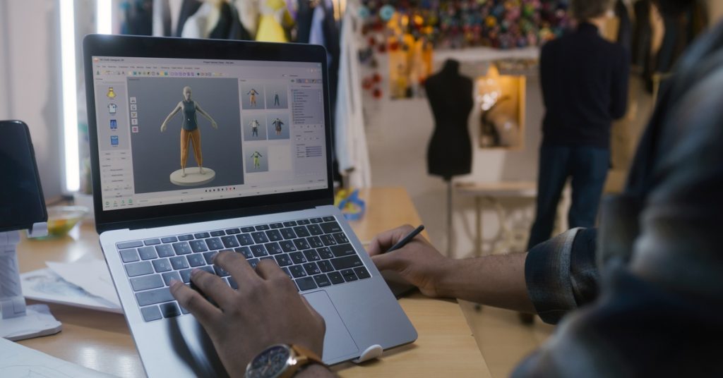 A man using a laptop that's propped up on a stand. He's looking at some digitized clothing while writing something down.