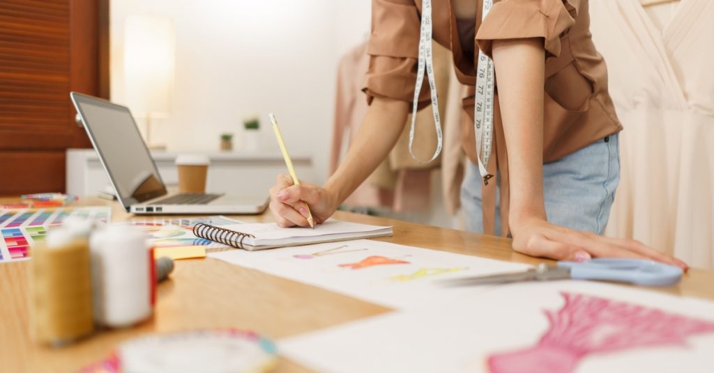 A woman leaning against a table with one hand while writing with the other. She is looking at pictures of dress designs.