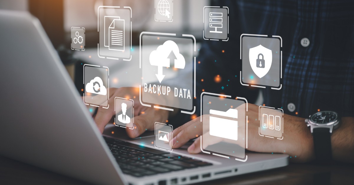 A man with a watch working at a laptop. There is a graphic above the keyboard with multiple data backup symbols.