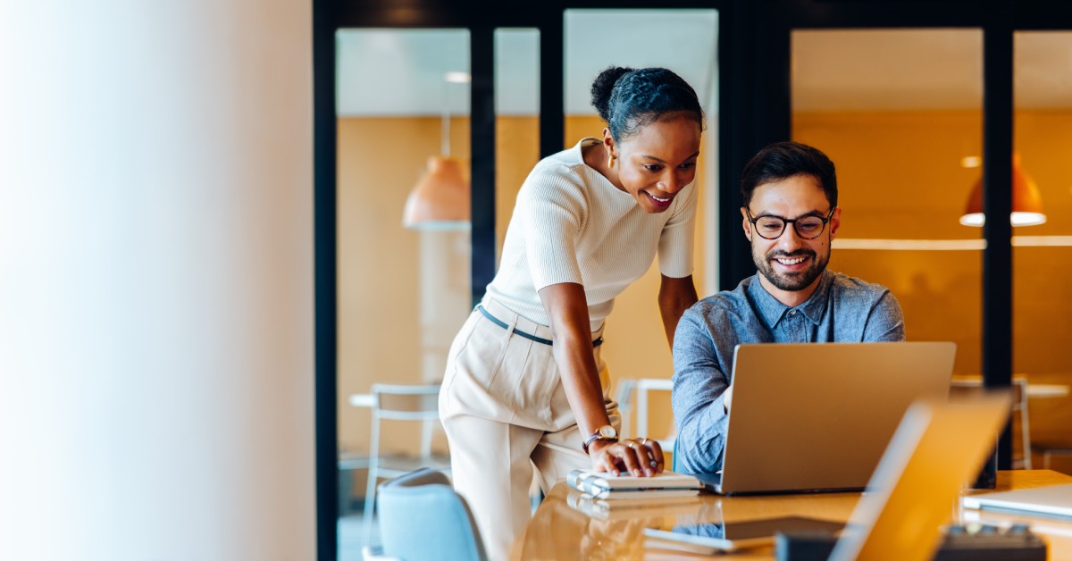 A man sitting at a laptop while a woman looks over his shoulder at what he's doing. They appear to be working together.