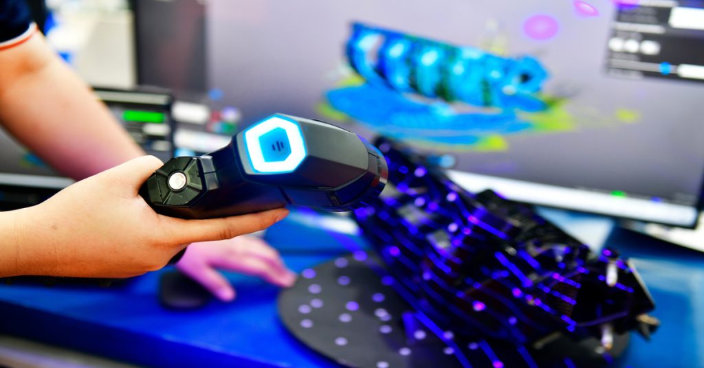 Someone using a portable 3D laser scanner on a metal object. The object has a render showing up on the computer screen.